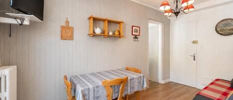 A dining area with a table and chairs, a television mounted on the wall, and a wooden shelf with kitchen items.