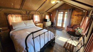 A rustic bedroom with a metal bed frame, wooden walls, a small table with a vase of flowers, and a chair.