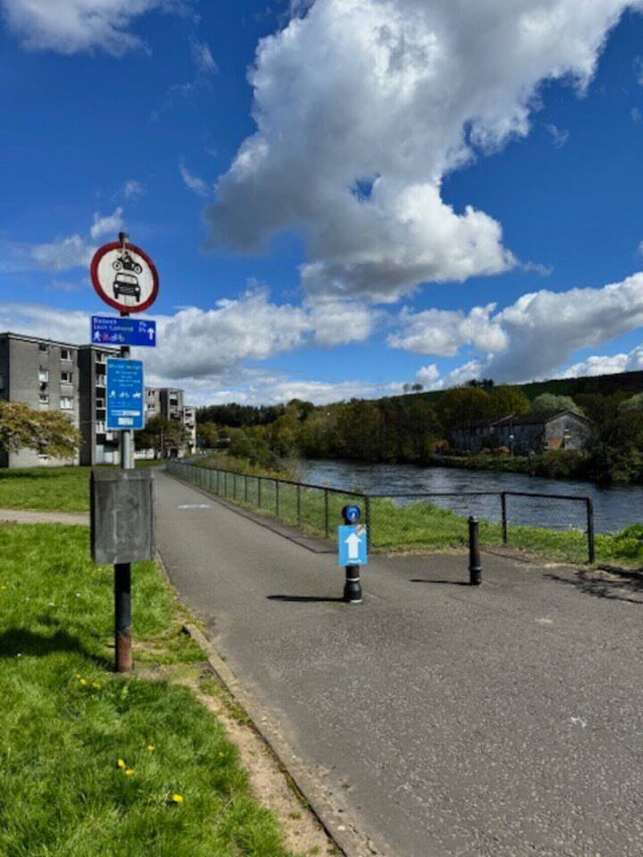 Un sendero junto a un río, con un cartel de prohibición de vehículos, un indicador direccional y una señal azul que marca una ruta peatonal.