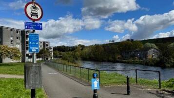 Un sendero junto a un río, con un cartel de prohibición de vehículos, un indicador direccional y una señal azul que marca una ruta peatonal.