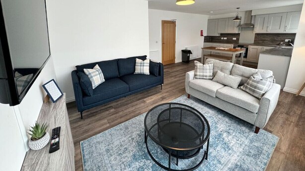 Una sala de estar moderna con un sofá azul, un sofá gris, una mesa de centro redonda y una cocina al fondo.