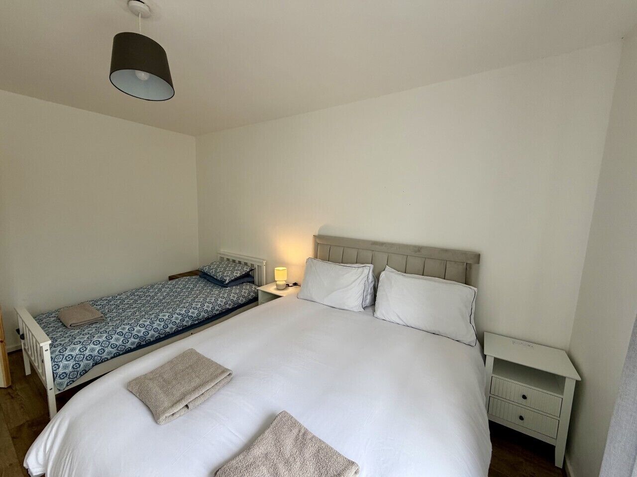 A single bed with a white comforter and two pillows, a bedside table with a lamp, and a small nightstand with drawers.