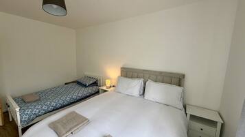 A single bed with a white comforter and two pillows, a bedside table with a lamp, and a small nightstand with drawers.