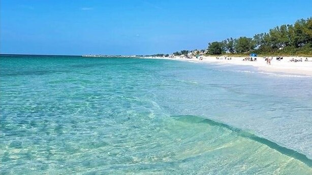 Une plage à l’eau turquoise et limpide, avec du sable blanc, quelques personnes sur le rivage et une rangée d’arbres en arrière-plan.