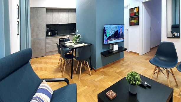 A modern living room with a sofa, a coffee table with a plant, a TV on the wall, and a dining area with a kitchen in the background. - Apart in the Heart Of Athens (Athina)