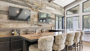 Una cucina moderna con parete in legno, un ampio televisore a schermo piatto, un angolo bar con sgabelli e una grande finestra vista alberi.