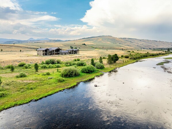 A two-story house with a large front porch and a garage, surrounded by a grassy field and a river.