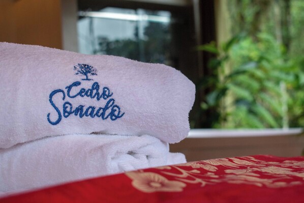 Toalla blanca doblada con cuidado, con texto bordado y un logotipo de árbol, sobre una cama con edredón rojo estampado con flores.