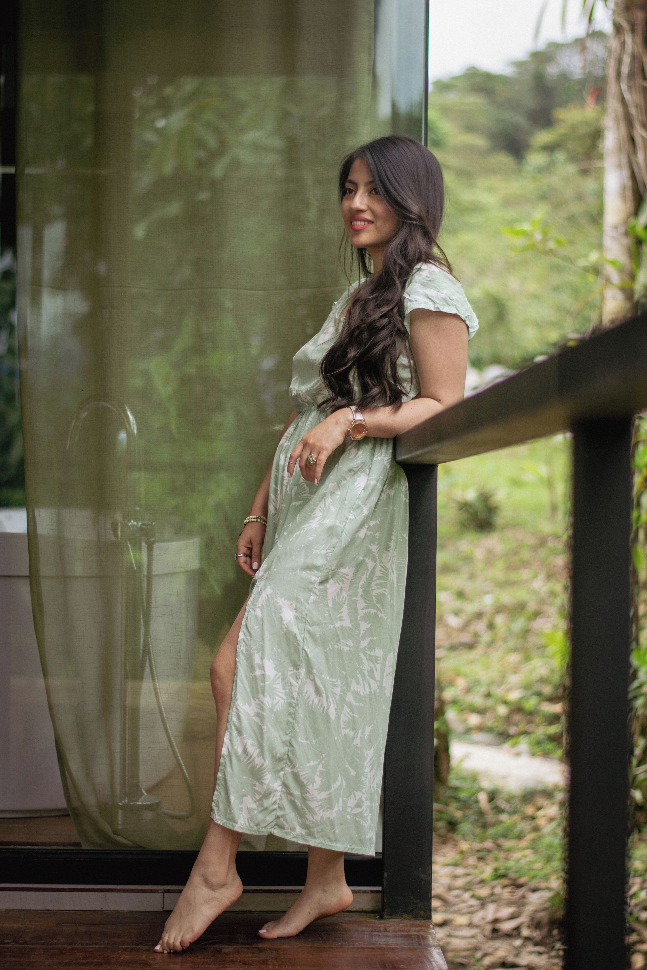 A woman in a light green dress leaning on a glass railing with a natural outdoor view.