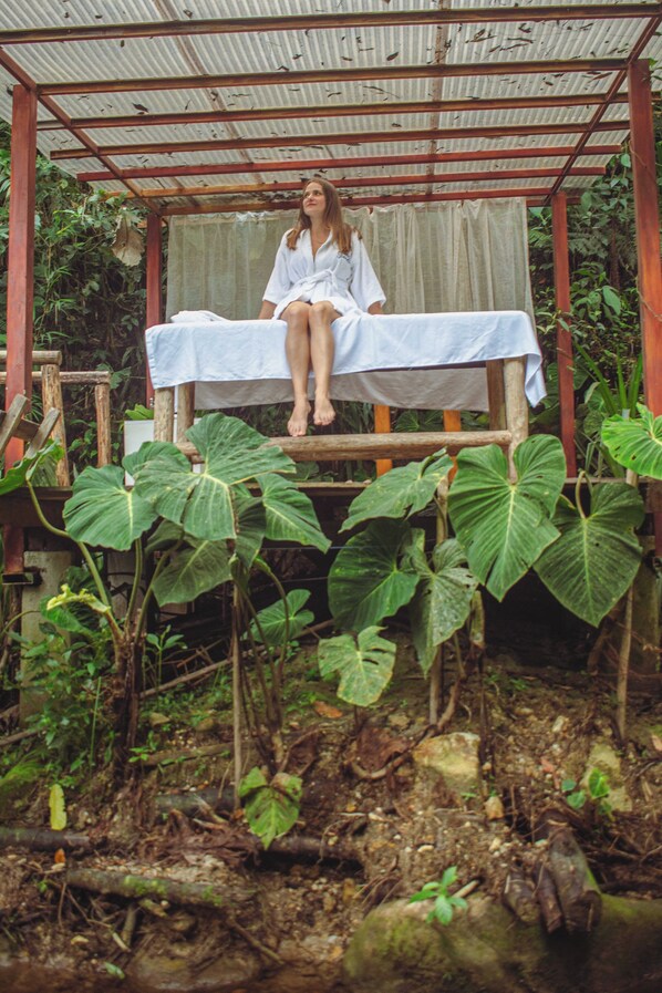 A person sitting on a wooden bench with a white cloth, surrounded by lush greenery and a metal roof structure.