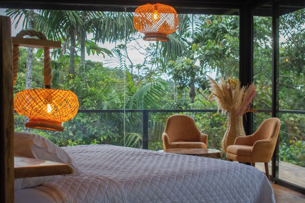 A bedroom with a large bed, two chairs, a small table, and a view of lush greenery through large windows.