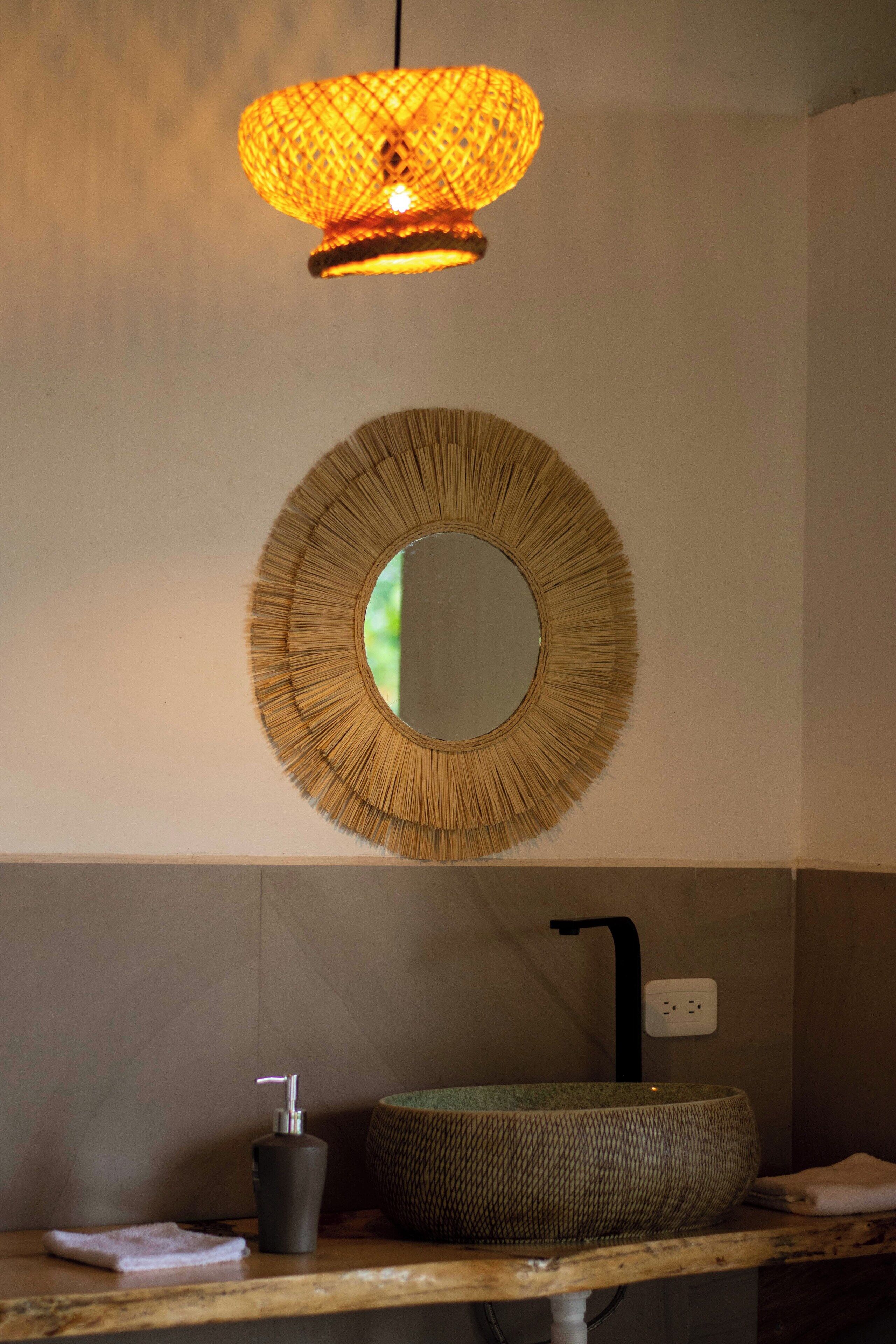 A bathroom with a round mirror, a wicker sink, a bottle of soap, and a hanging light fixture.