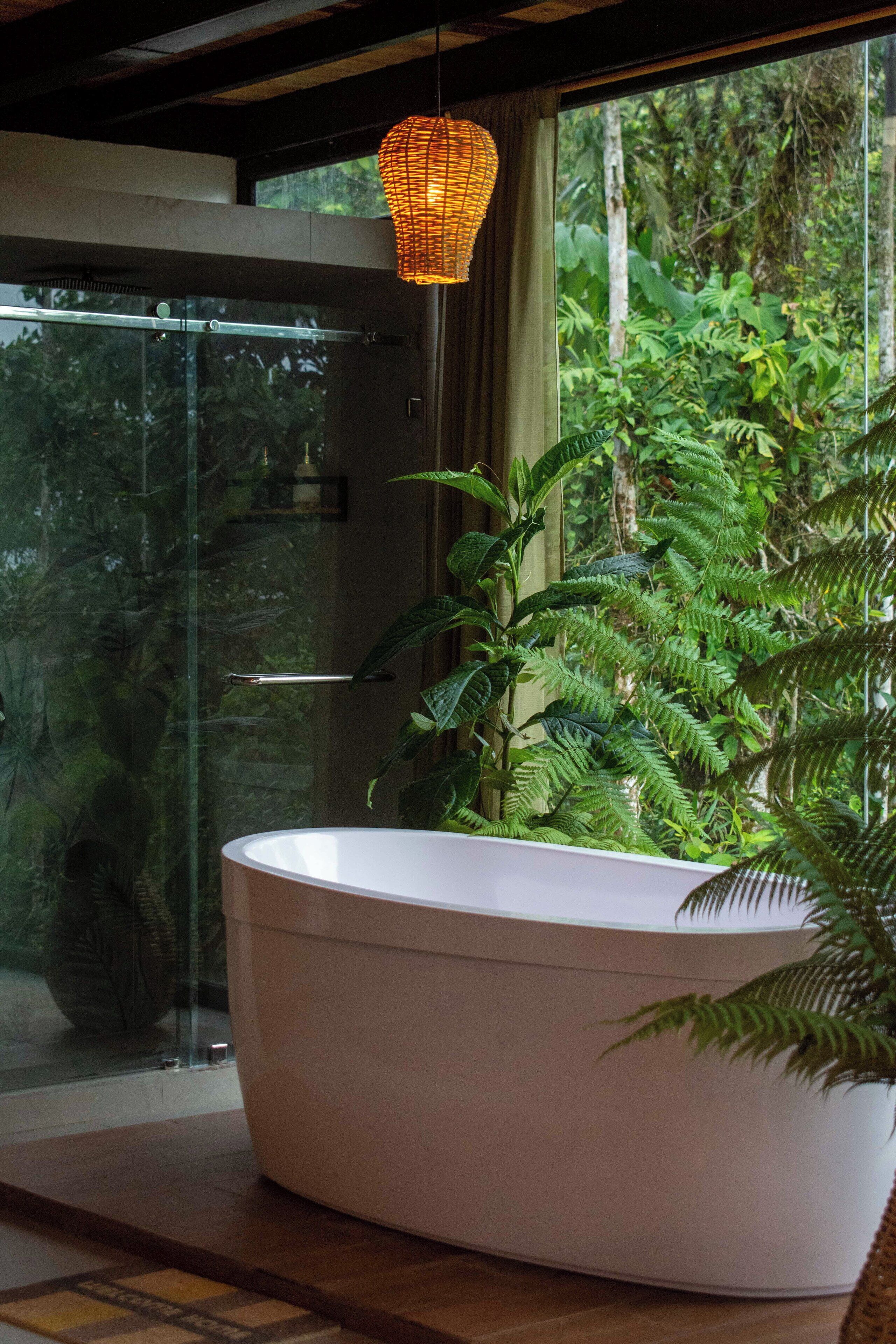A modern bathroom with a large white bathtub, a glass shower enclosure, and lush greenery visible through the window.