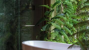 A modern bathroom with a large white bathtub, a glass shower enclosure, and lush greenery visible through the window.