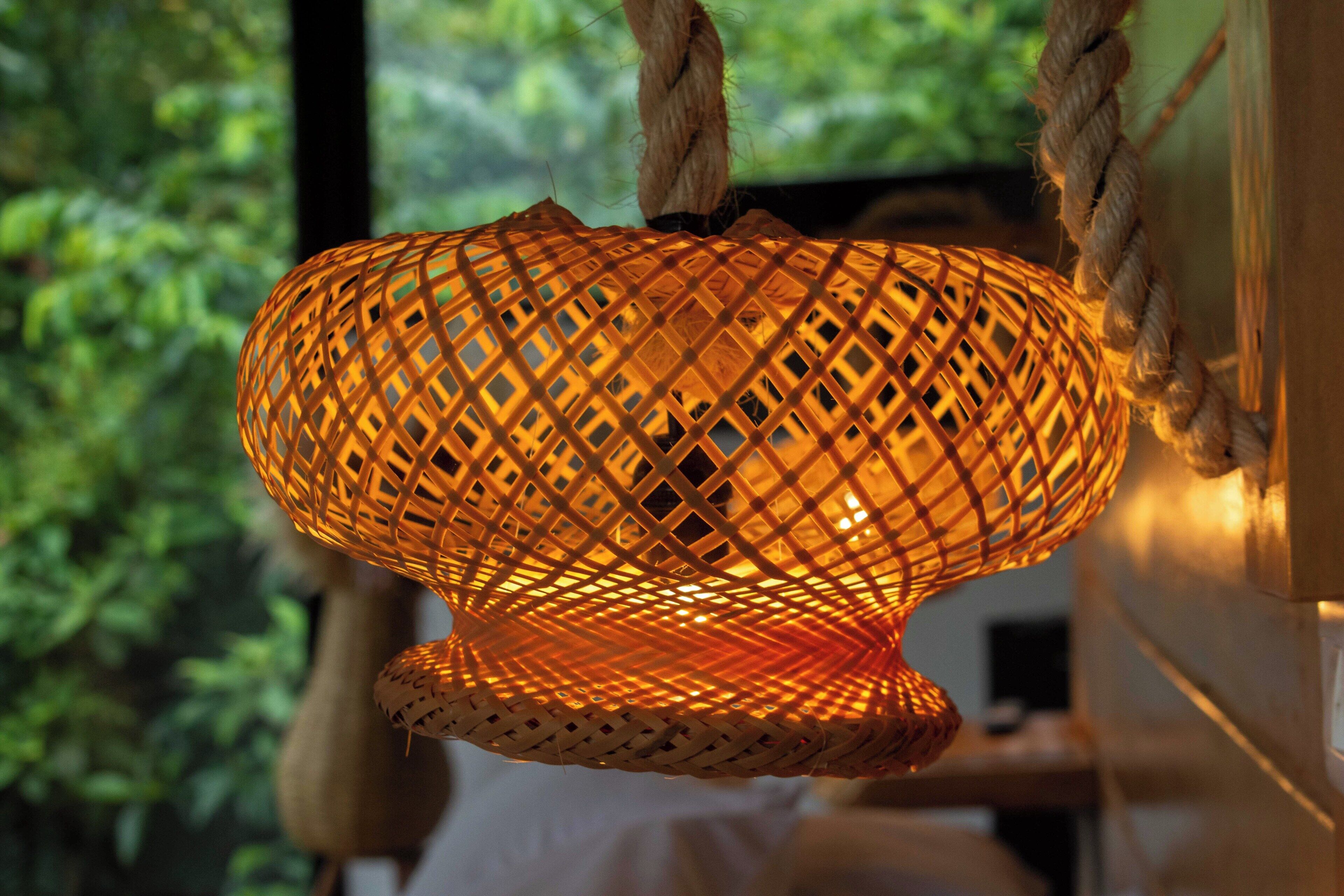 A woven hanging light fixture with a warm glow, suspended by ropes, in a room with a view of greenery outside.