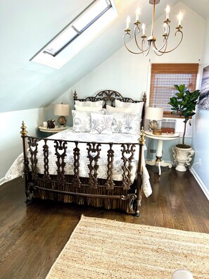 A bedroom with a sloped ceiling, a bed with ornate metal railings, a chandelier, a window with blinds, and a potted plant. - Bernie Beach House And Bungalow  (Lavallette)