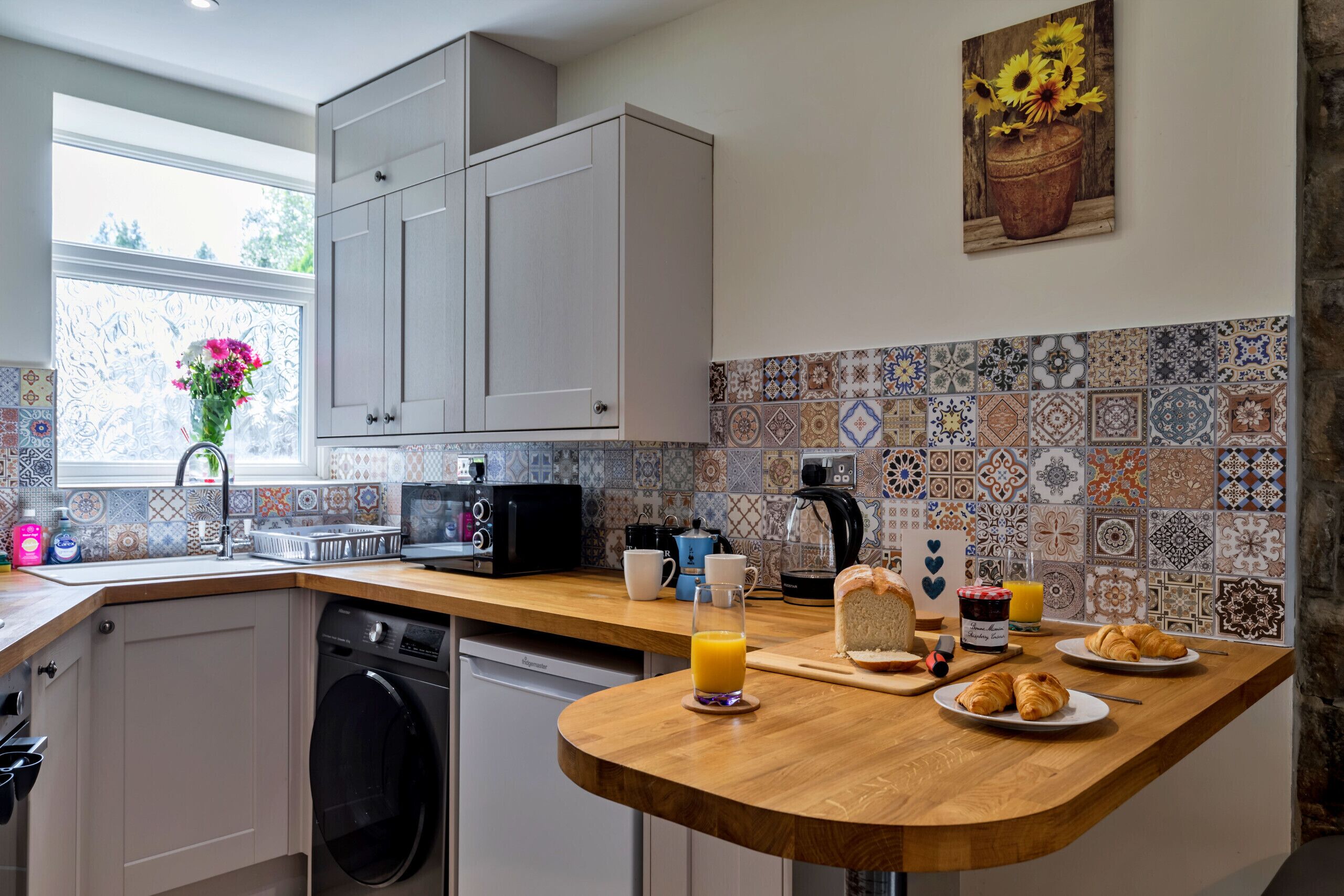 A kitchen with a wooden countertop, a washing machine, a coffee maker, and a breakfast setup with croissants and orange juice.