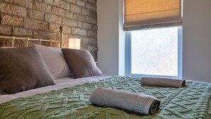 Ein Schlafzimmer mit einer Ziegelwand, einem Bett mit einer grün gemusterten Tagesdecke, zwei Kissen und einem aufgerollten Handtuch auf dem Bett.