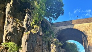 Eine Steinbogenbrücke über einer felsigen Klippe, im Vordergrund Gras.