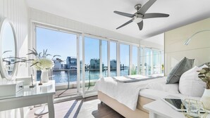 A modern bedroom with a large window overlooking a body of water, a ceiling fan, and a bedside table with a tablet.