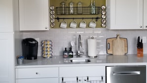 Eine Küche mit weißen Schränken, einem Edelstahl-Geschirrspüler, einer Kaffeemaschine, einem Messerblock und einem hängenden Regal mit Pflanzen.