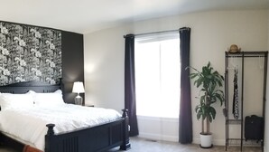 Ein Schlafzimmer mit einem großen Bett, einem Nachttisch, einer Lampe, einem Blumentopf, einer Heizung und einem Fenster mit Vorhängen.