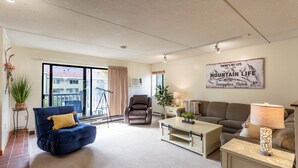 Un salon moderne avec un fauteuil bleu, un fauteuil en cuir marron, un canapé et une table basse. On y trouve une grande fenêtre avec des rideaux, une plante et un panneau décoratif fixé au mur.