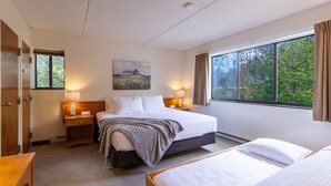 Une chambre d’hôtel avec un grand lit, un lit plus petit, une table de chevet avec une lampe, une armoire en bois et une fenêtre donnant sur des arbres.