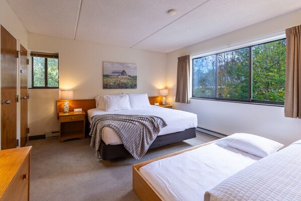 A hotel room with a large bed, a smaller bed, a nightstand with a lamp, a wooden wardrobe, and a window with a view of trees.