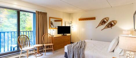 Ein Hotelzimmer mit einem Bett, einem Schreibtisch mit Fernseher, einem Sessel, einer Kommode und einem Balkon mit Blick auf Bäume.