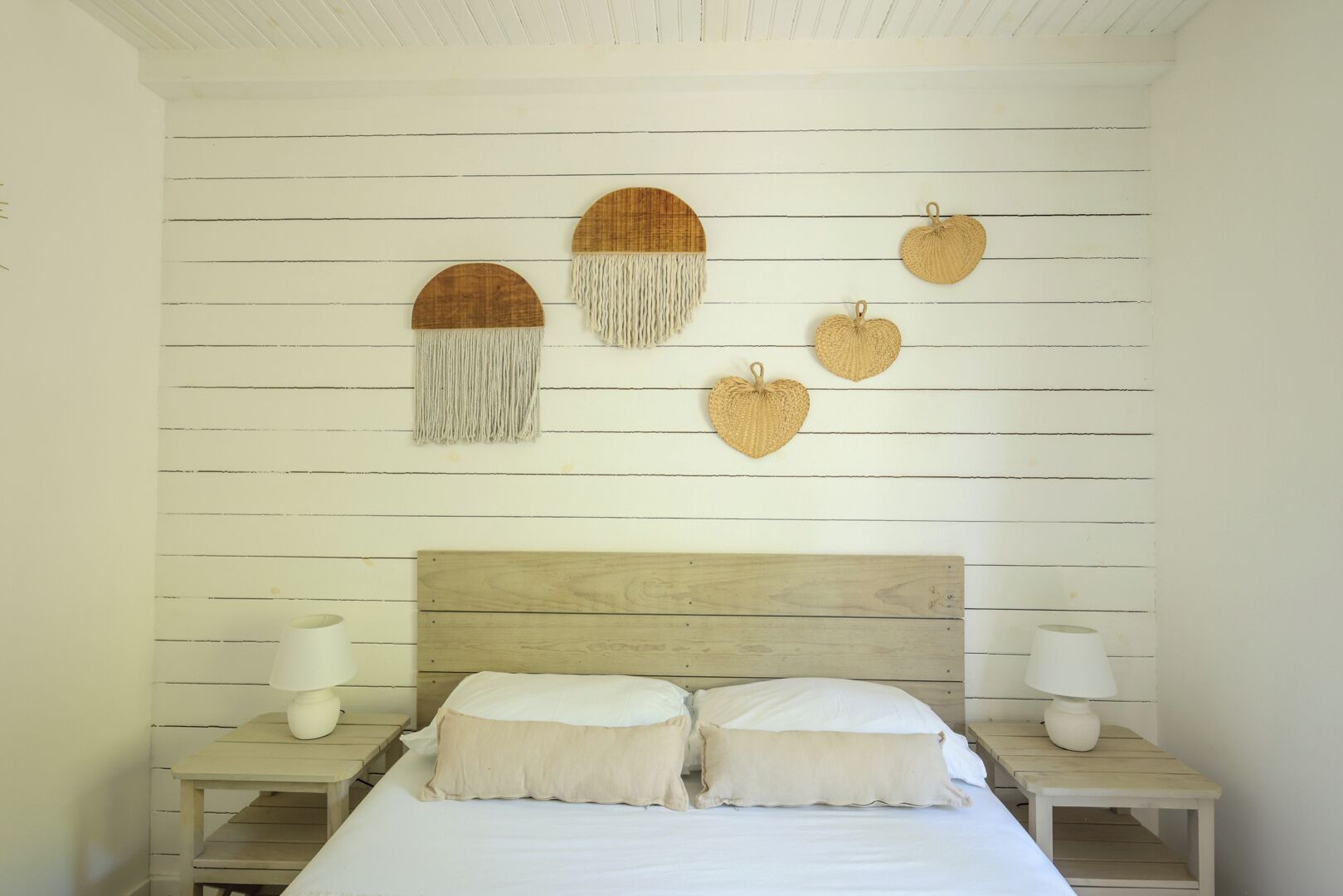 A bedroom with a wooden headboard, a bed with two pillows, and wall-mounted decorative items including a circular wooden piece with tassels and several heart-shaped items.