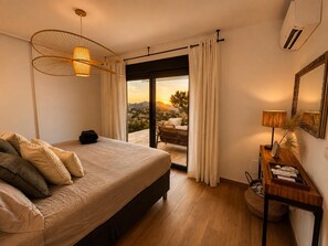 Un dormitorio con cama, mesita de noche, lámpara, espejo y vista al exterior.