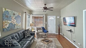 Ein Wohnzimmer mit einem schwarzen Ledersofa, einem gemusterten Hocker und einem an der Wand befestigten Fernseher.