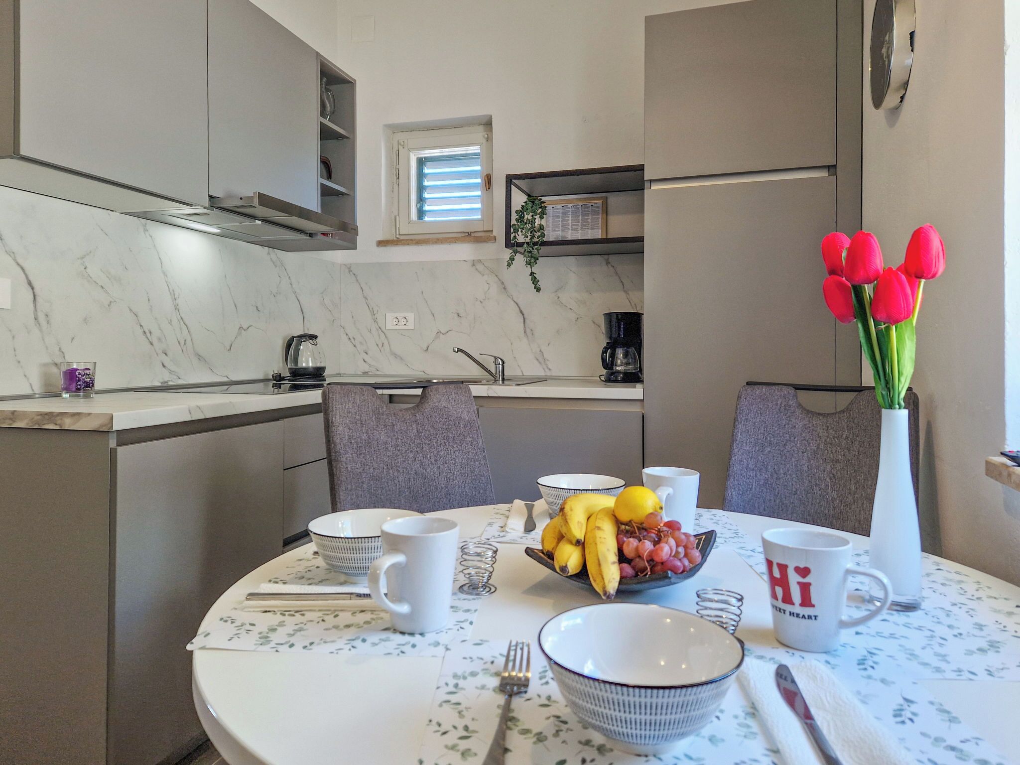 A modern kitchen with a marble countertop, a small window, and a dining table set with a bowl of fruit, a coffee maker, and a vase of tulips.
