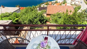 A balcony with a table set for two, overlooking a coastal town with terracotta-roofed buildings and a view of the sea.