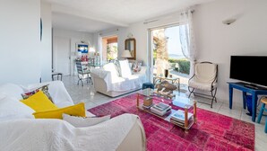 Een ruime woonkamer met een grote bank, een salontafel en een televisie. Er is een eethoek met stoelen en een tafel, en een balkon met uitzicht.