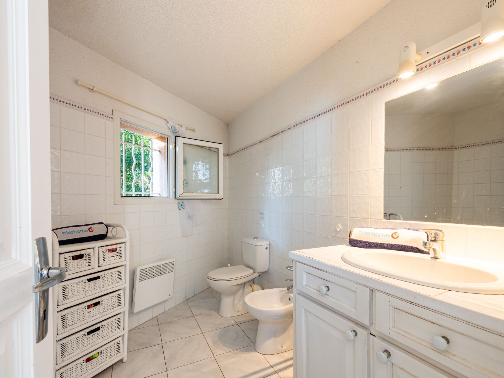 Une salle de bain comprenant des toilettes, un bidet, des murs recouverts de carreaux blancs, une fenêtre et un lavabo blanc avec un miroir.