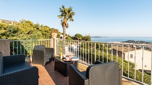 Een balkon met rieten meubilair, een palmboom en uitzicht op zee en gebouwen.
