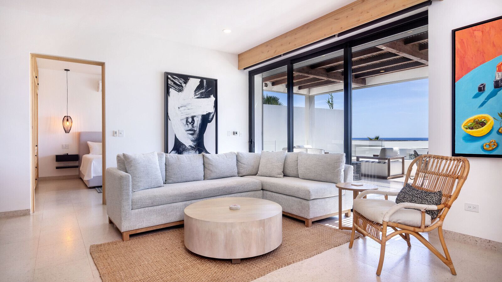 A modern living room with a grey sectional sofa, a round coffee table, and a wicker chair. There is a large window with a view of the sea.