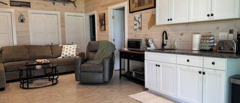 A cozy living room with a sofa and armchair, a small coffee table, and a kitchen area with white cabinets and a microwave.