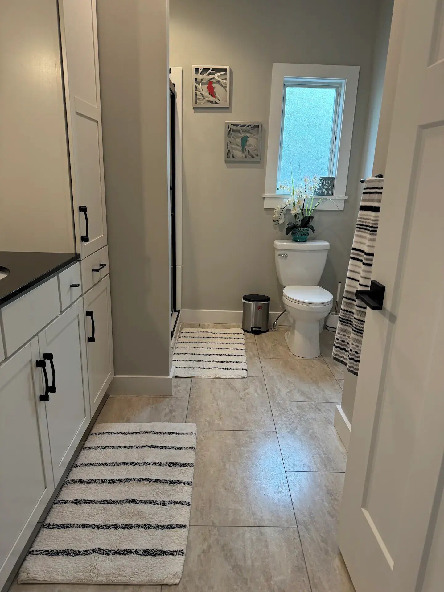 Une petite salle de bain comprenant des toilettes, un lavabo et des placards. On y trouve deux tapis, une fenêtre au-dessus des toilettes et une petite plante en pot.
