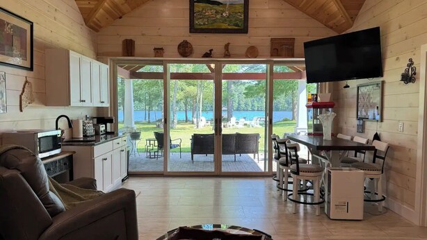 Interior acogedor de una cabaña con comedor, cocina y un amplio ventanal corredera que da a un jardín trasero con vista a un lago.