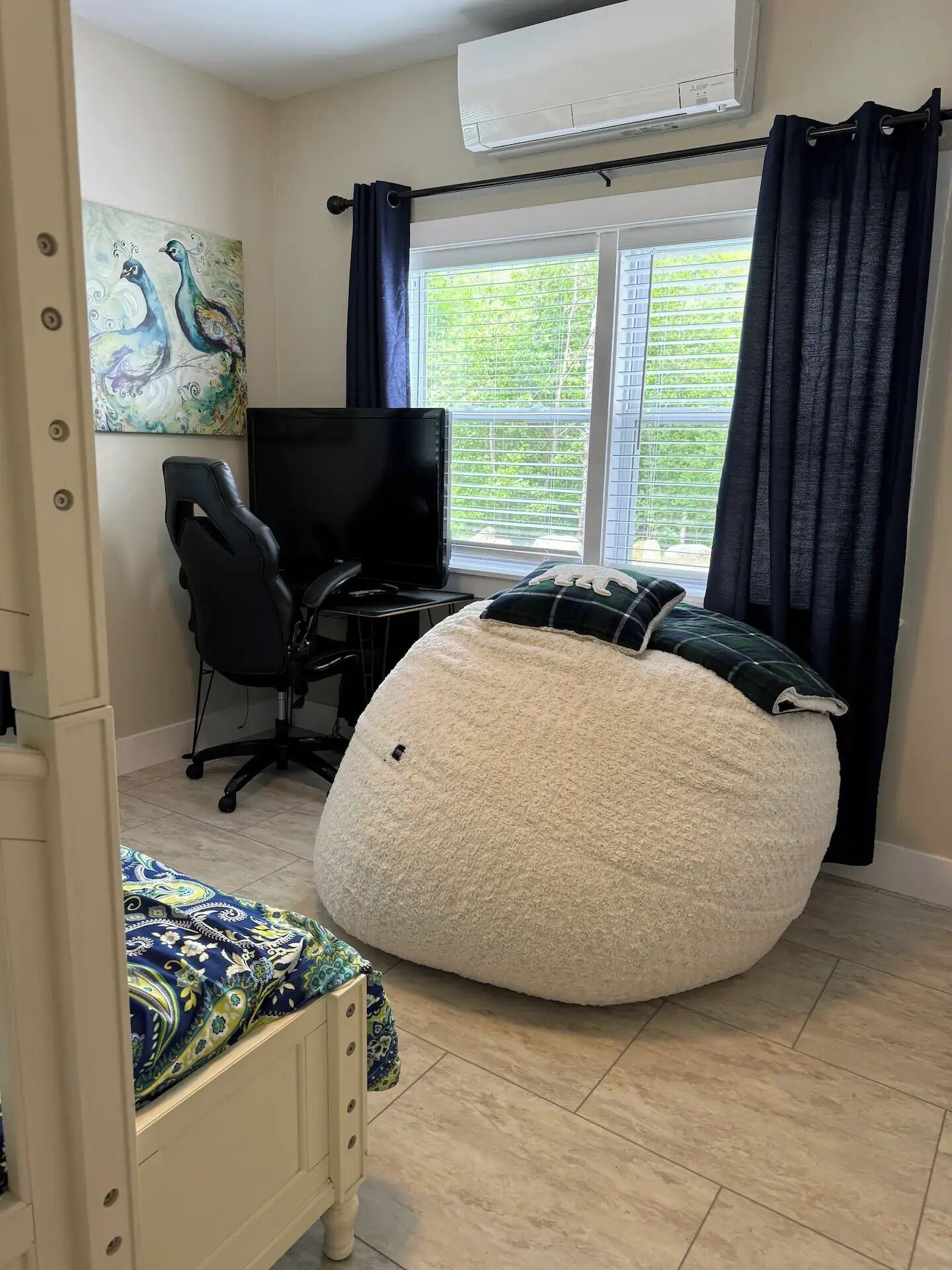 Une pièce comprenant un fauteuil en forme de sac de haricots, un bureau avec un ordinateur, une chaise, un lit avec une couvre-lit à motifs, un tableau représentant des paons et une fenêtre avec des rideaux.