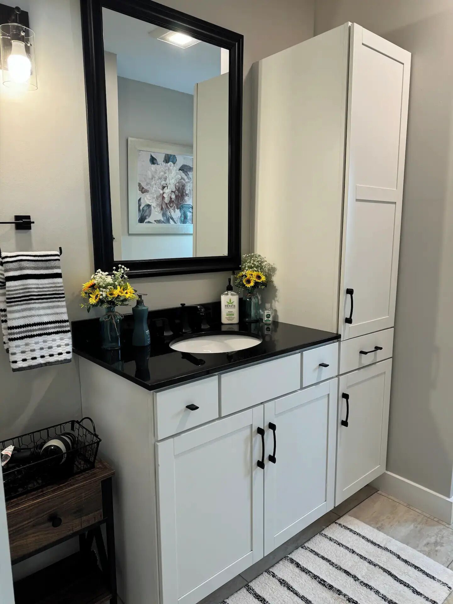 Une salle de bain moderne avec un lavabo sur un plan de toilette noir, des meubles blancs, un grand miroir et des fleurs décoratives.