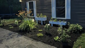 Una casa con revestimiento oscuro, dos ventanas con contraventanas blancas y un jardín con diversas plantas y un parterre elevado.