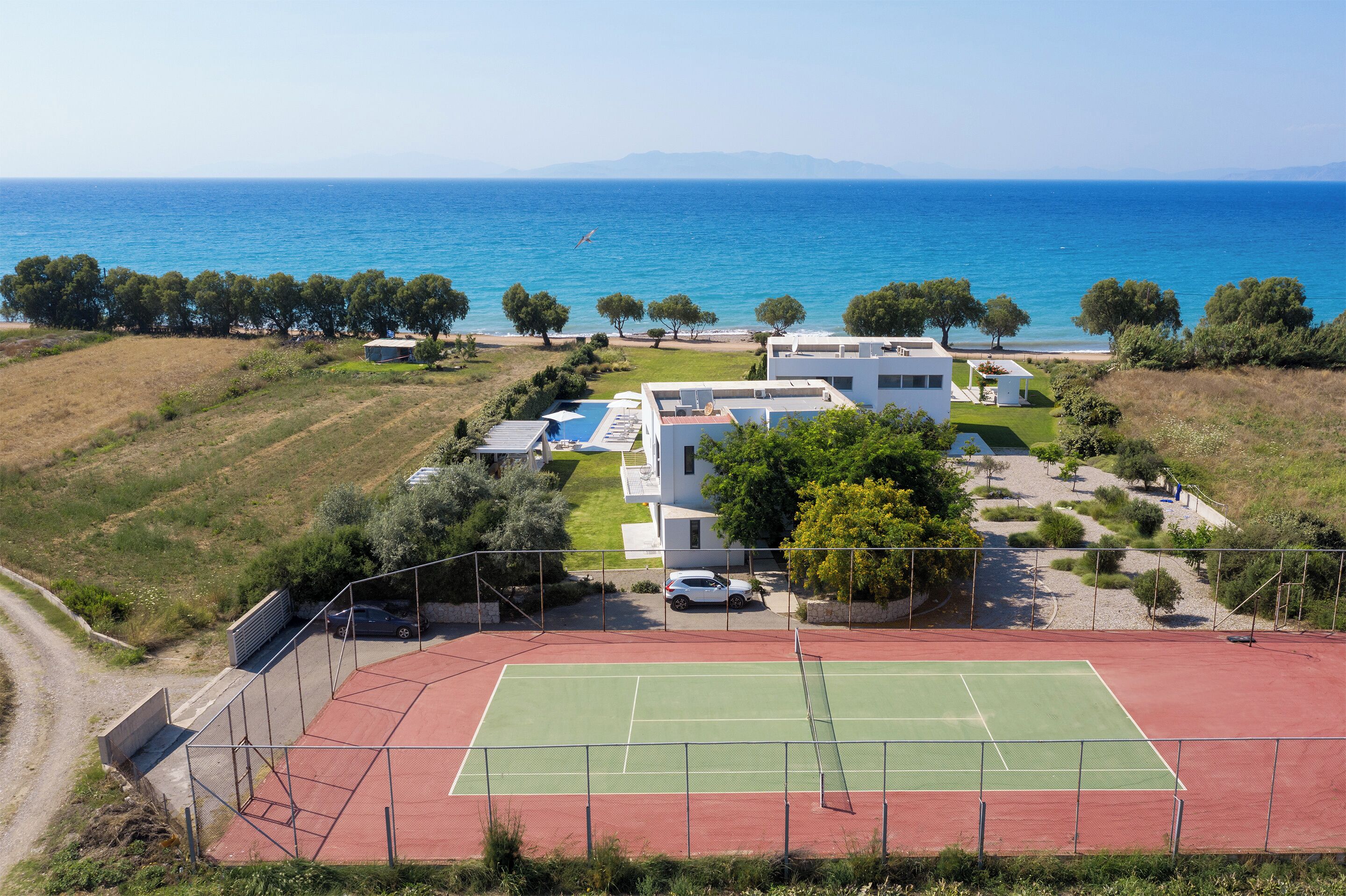 Un court de tennis entouré d’une zone délimitée par une clôture, d’un bâtiment blanc et offrant une vue sur la mer.