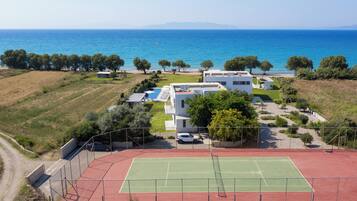 Un court de tennis entouré d’une zone délimitée par une clôture, d’un bâtiment blanc et offrant une vue sur la mer.