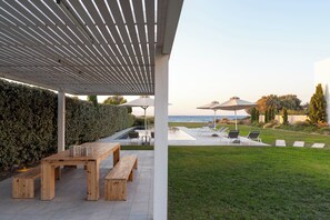 A covered outdoor area with a wooden table and benches, a pool, and lounge chairs with umbrellas.