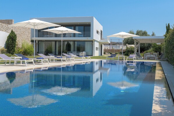 A modern pool area with lounge chairs and umbrellas, a stone wall, and a clear blue sky.