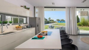 A modern kitchen with a central island, a built-in oven, and a view of a landscaped garden. - Stunning beachfront retreat, private tennis court & infinity pool (Rhodes)